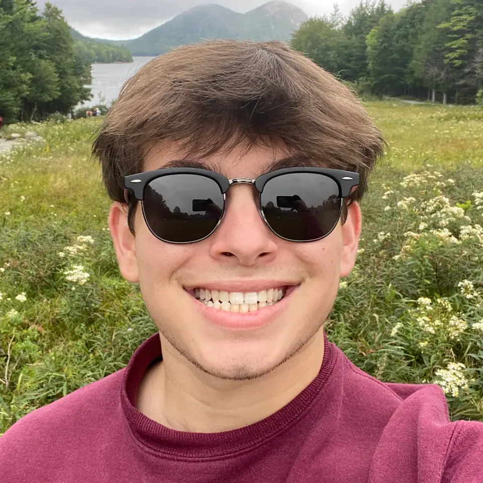 Corey smiling in sunglasses and a maroon sweatshirt, standing in a field of wildflowers with a lake and mountains behind him.